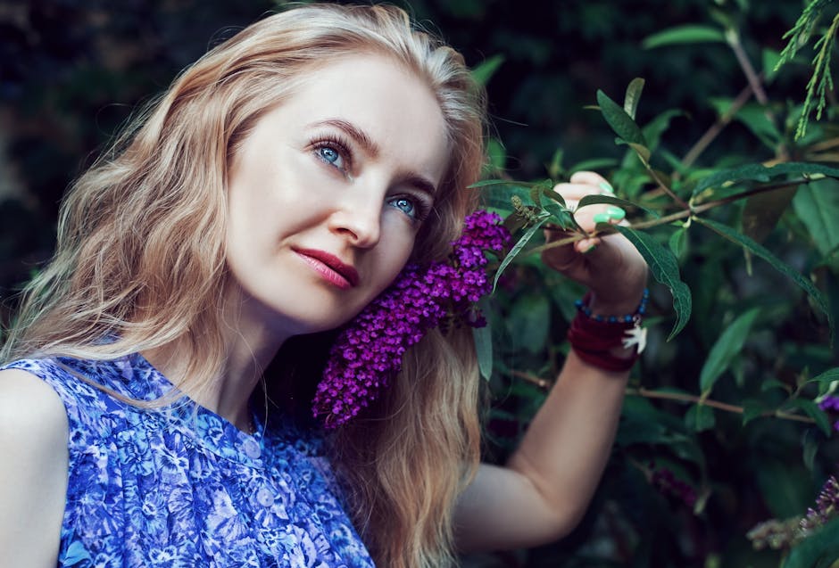 Blonde woman enjoying nature, posing with vibrant purple flowers in a lush outdoor setting.