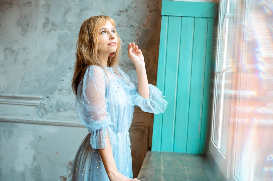 Woman in a sheer blue dress stands gracefully by a sunlit window indoors.