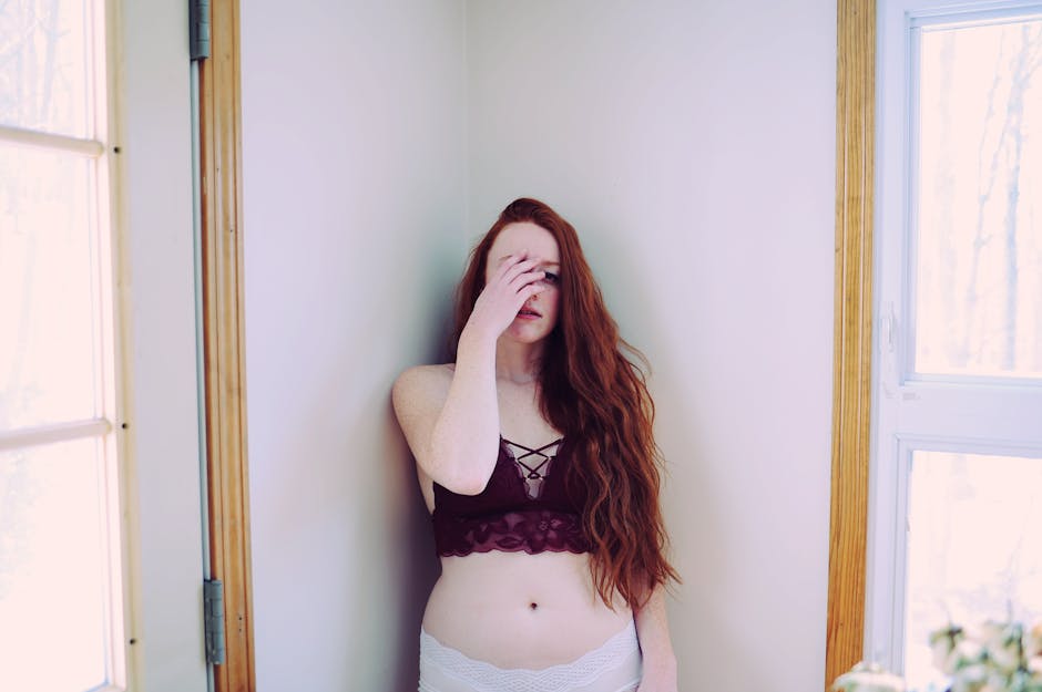 A young woman stands indoors, covering her face while wearing stylish lingerie, conveying emotion.