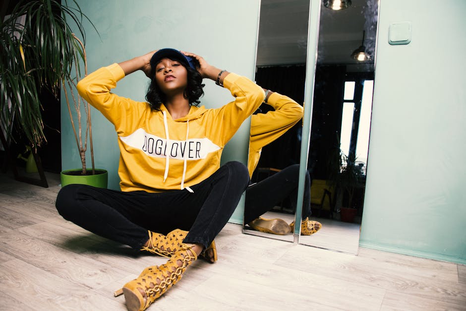 Young woman in a yellow hoodie posing casually indoors with a mirror reflection, expressing fashion and style.