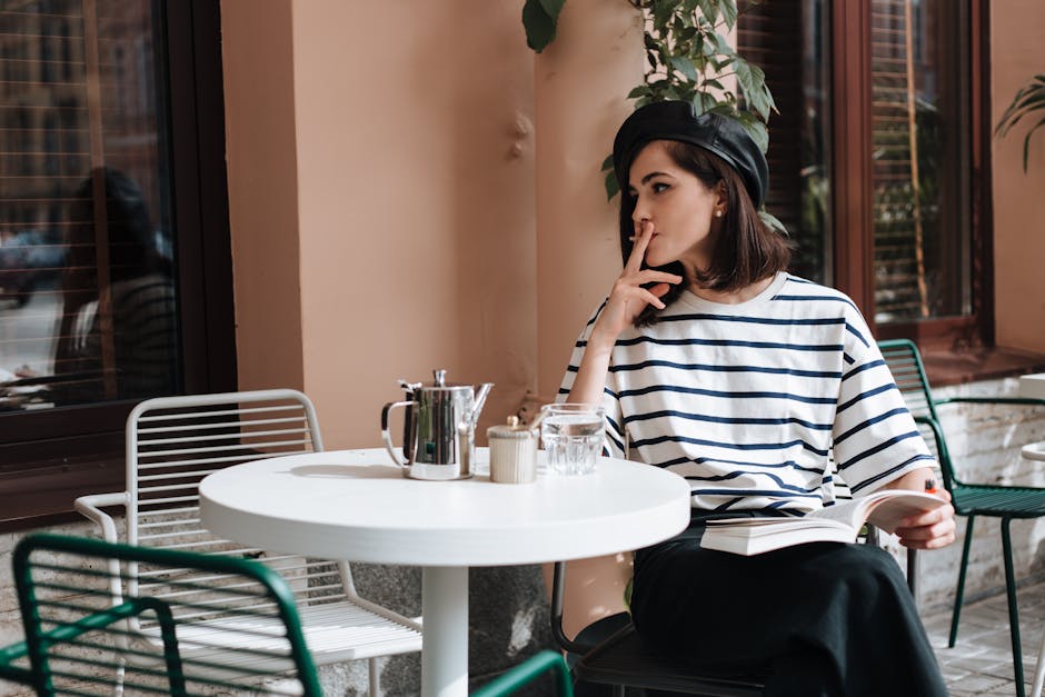 Woman in striped shirt sitting at an outdoor cafe, holding a book and enjoying a smoke.