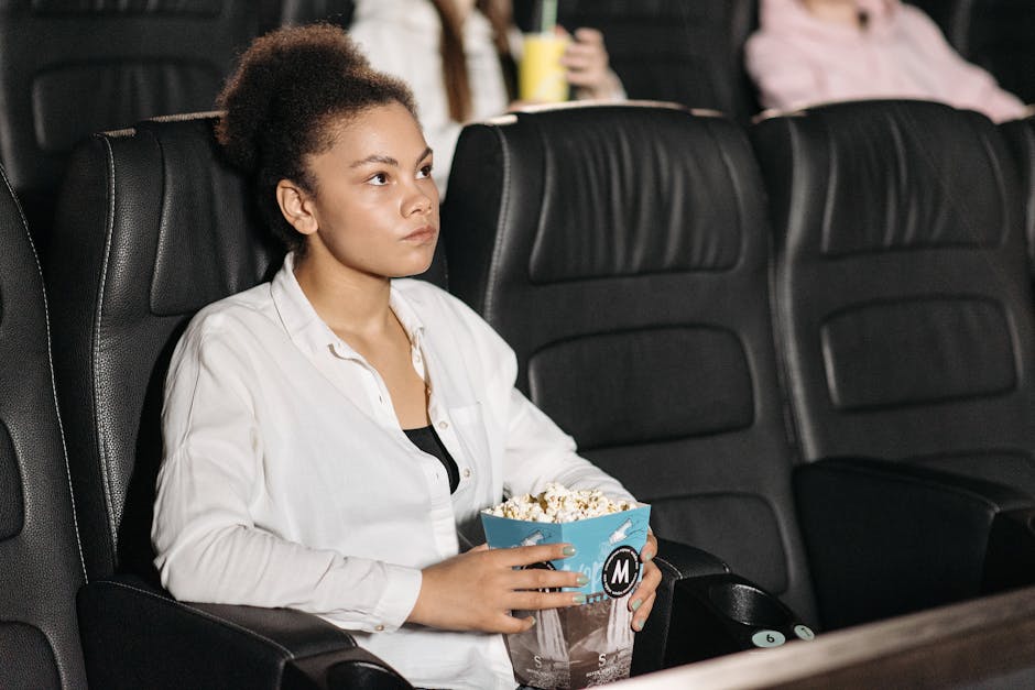 A woman sits in a movie theater holding popcorn, intently watching the screen.