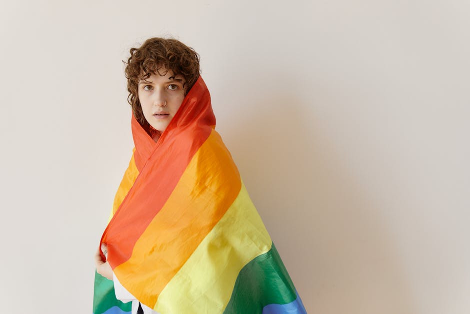 Young adult with curly hair wrapped in rainbow pride flag, celebrating LGBTQ+ identity.