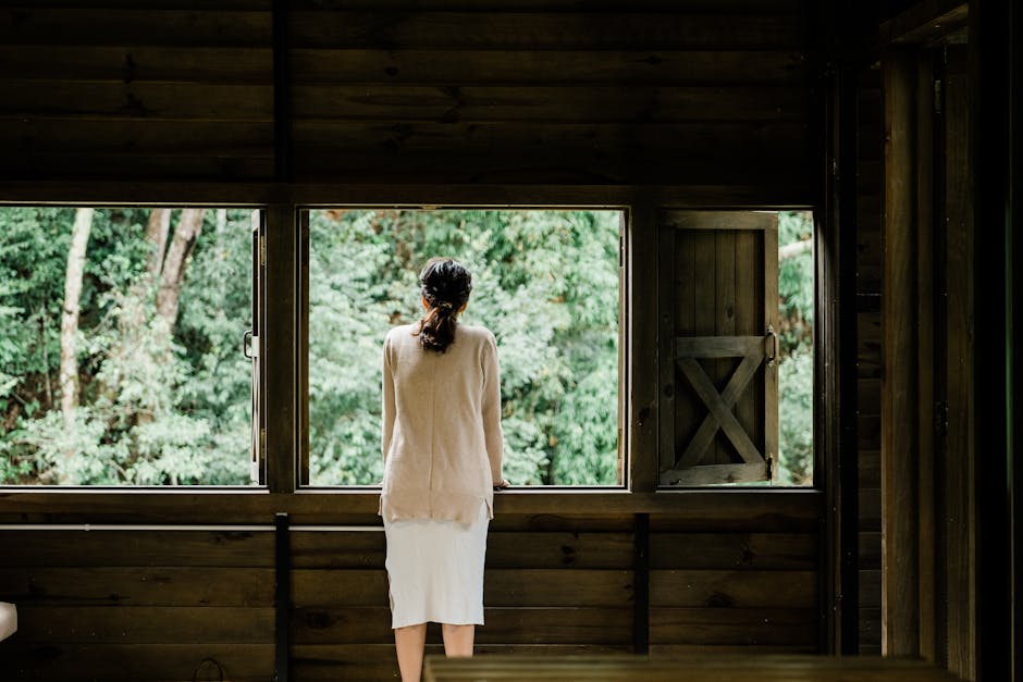A woman stands indoors, looking out a wooden window at a lush green forest.
