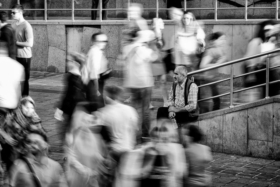An elderly man sitting amidst a blur of bustling city pedestrians.