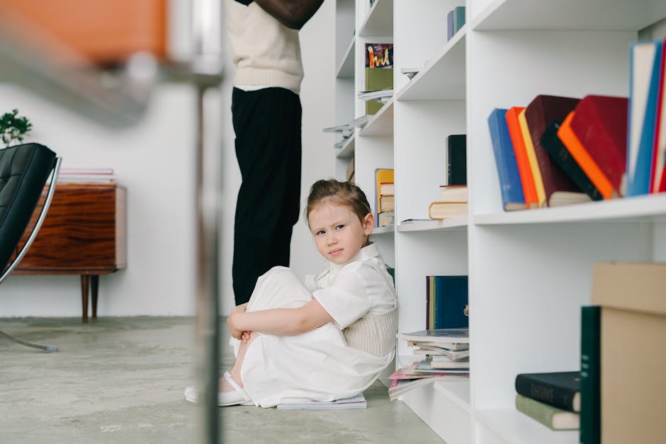A young girl in a white dress sitting by a bookshelf in a modern library setting.