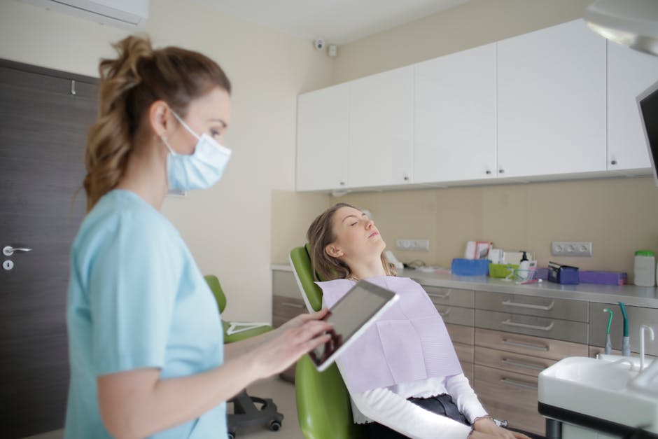 Female dentist in uniform and sterile mask touching tablet screen while standing near female patient sitting on medical chair