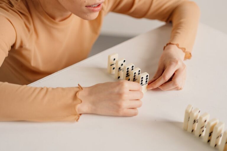 A woman in beige long sleeves setting up dominoes in a line on a white table.
