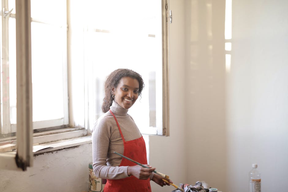 Young smiling lady in casual outfit and apron standing near bright window and preparing for work with paintbrushes in hands