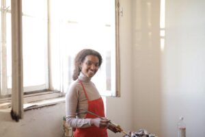 Young smiling lady in casual outfit and apron standing near bright window and preparing for work with paintbrushes in hands