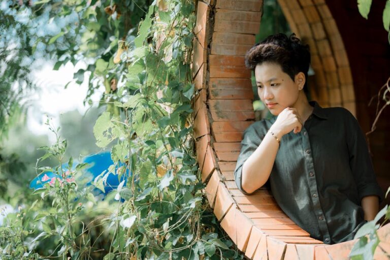 A person in casual attire sits thoughtfully in a brick arch surrounded by lush green vines.
