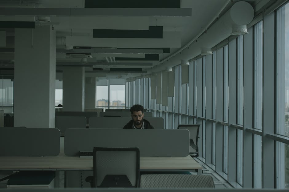 An adult man works alone in a contemporary open-plan office, surrounded by desks and dividers.