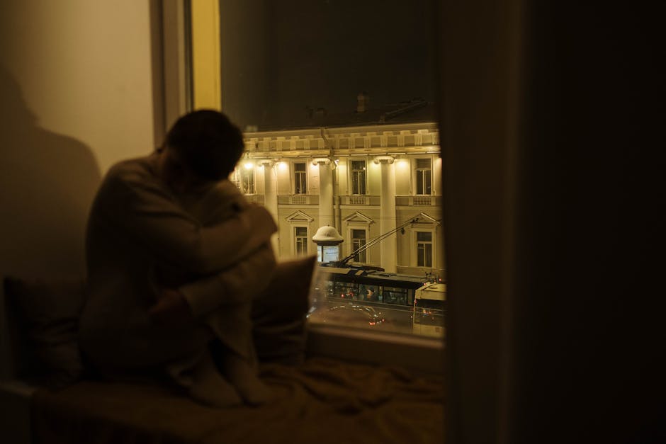 A pensive person sitting by the window at night overlooking a street with historical buildings.