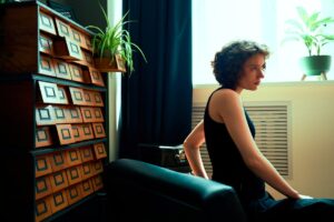 Pensive young woman seated by window in cozy room with plants and vintage decor.