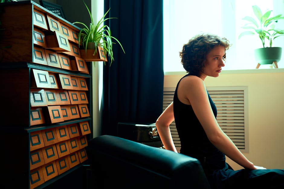 Pensive young woman seated by window in cozy room with plants and vintage decor.