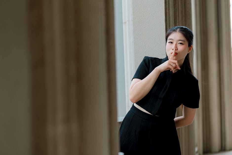 A young woman in black gestures for silence in a light-filled corridor, creating intrigue.