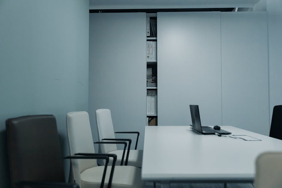 A sleek conference room with a white table and chairs, featuring a laptop on the table.