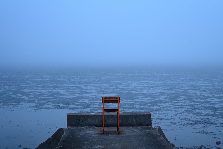 A solitary orange chair on a concrete pier overlooking a foggy ocean creates a serene, moody scene.