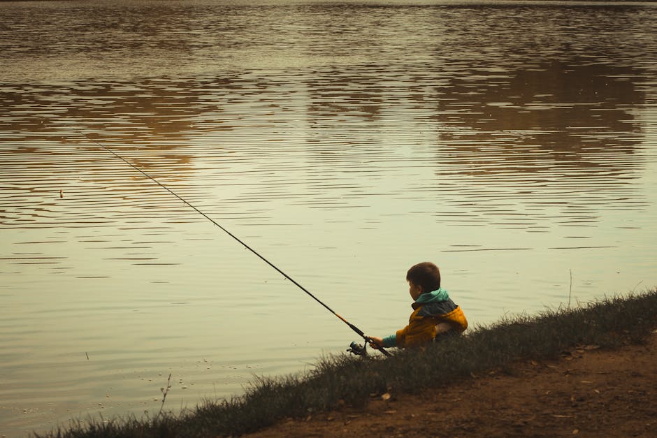 A young boy in a yellow jacket fishing by a serene lake in Hungary during a calm day.