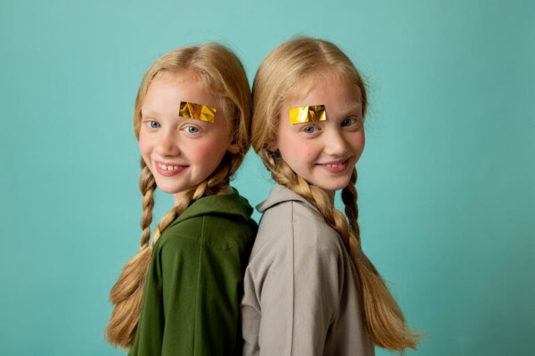 Adorable twin sisters with braids and gold face stickers, posing back-to-back indoors against a turquoise background.