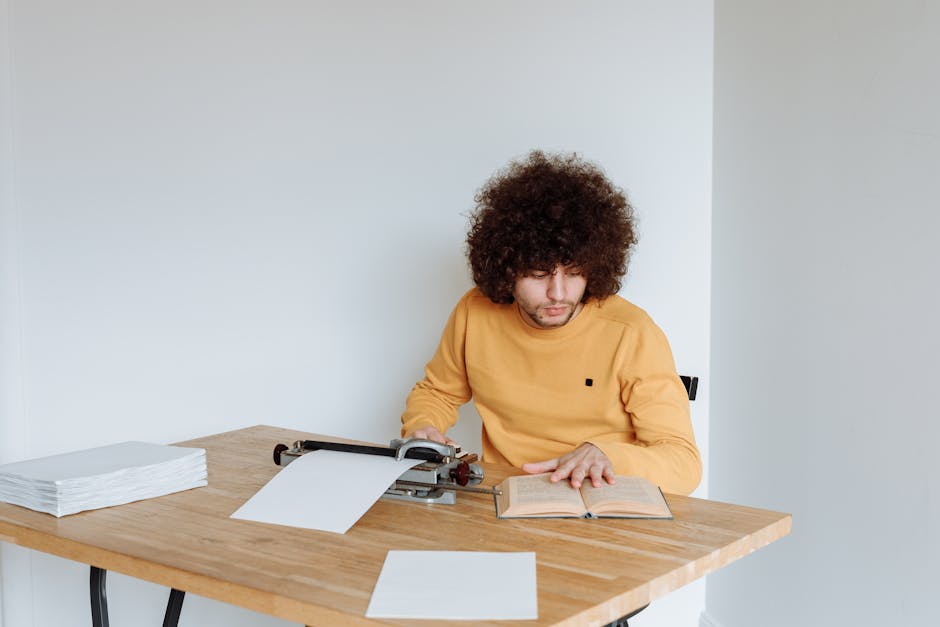 Young adult reading a book beside a typewriter, focused at a wooden table indoors.