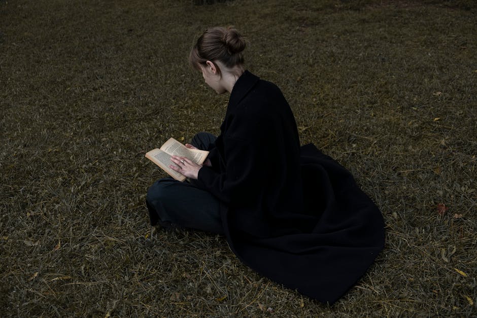 A woman sitting on grass reads a book. Captured from behind in a black coat.