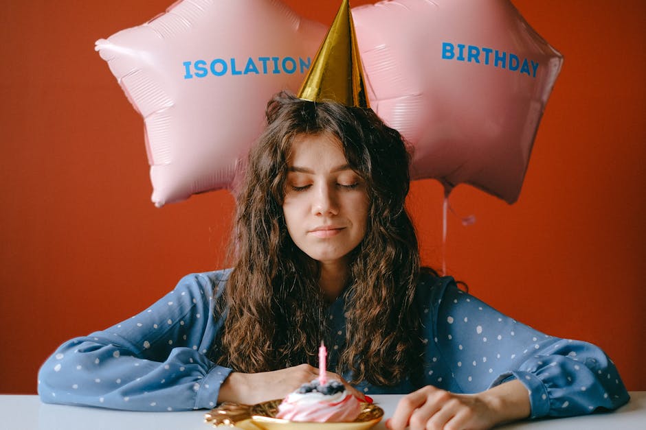 A woman in a party hat celebrates her birthday in isolation with balloons and a cupcake.