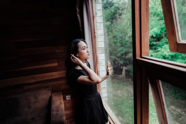 A thoughtful woman gazes outside from a wooden interior space, surrounded by nature.