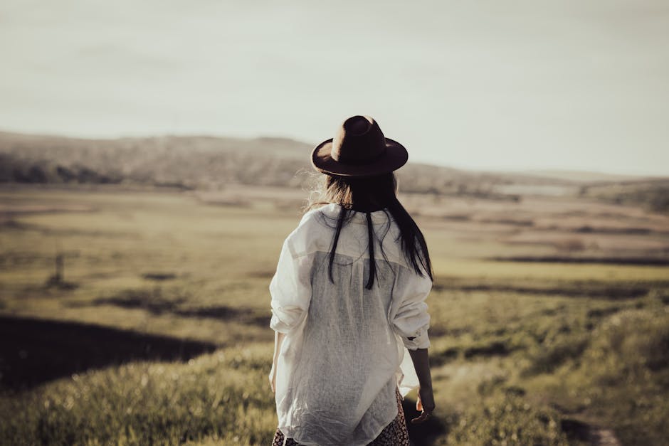 A woman with a hat stands in a vast field under the warm sunlight, embracing freedom.