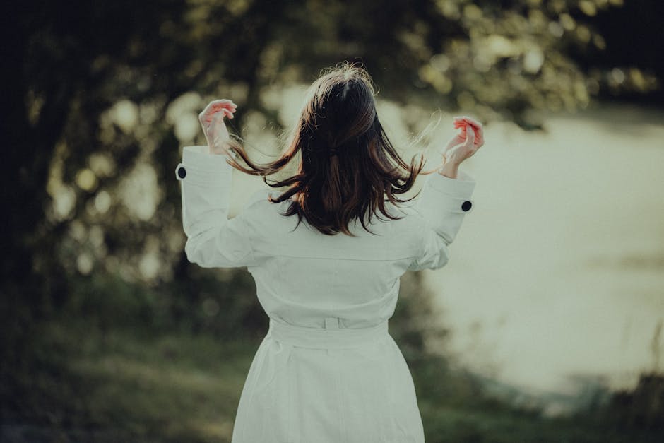 A woman in a white trench coat adjusts her hair in a sunny park setting.
