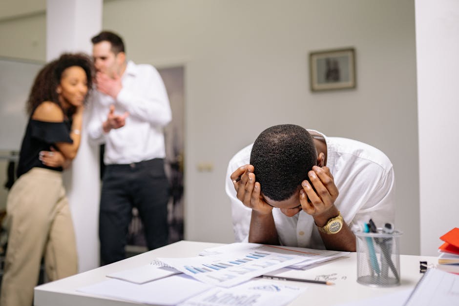 Despairing worker at office desk as colleagues gossip in background illustrating workplace bullying.